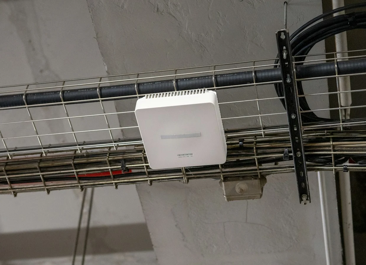 A white Wi-Fi access point mounted on a cable tray under an office ceiling