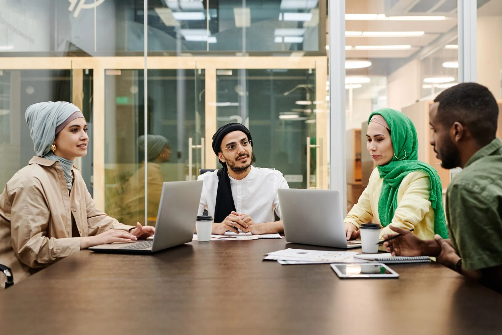Four colleagues with laptops around a table in a glass-walled meeting room